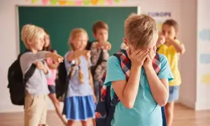 Smutny chłopiec zakrywa twarz, podczas gdy inne dzieci wskazują na niego palcami. To obraz tego, co zrobić, gdy dziecko jest wyśmiewane w szkole.