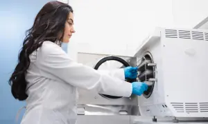 Kobieta w białym fartuchu i niebieskich rękawiczkach wkłada tacki do autoklawu. Technik sterylizacji medycznej - czy warto?