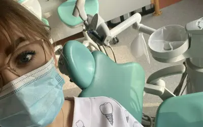 Praktyki asystentka stomatologiczna w gabinecie. Widoczny fragment twarzy w maseczce, okulary i fartuch z motywem zębów.