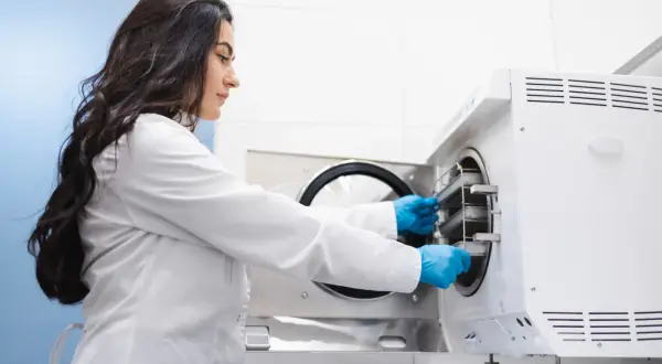 Kobieta w białym fartuchu i niebieskich rękawiczkach wkłada tacki do autoklawu. Technik sterylizacji medycznej - czy warto?