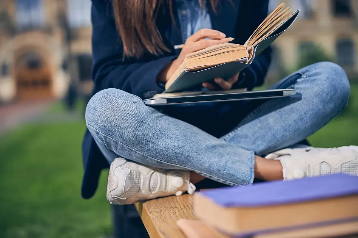 Dziewczyna w jeansach i marynarce uczy się na ławce, trzymając otwartą książkę i notując. To idealny przykład, jak można połączyć naukę w szkole zaocznej co to z życiem codziennym.