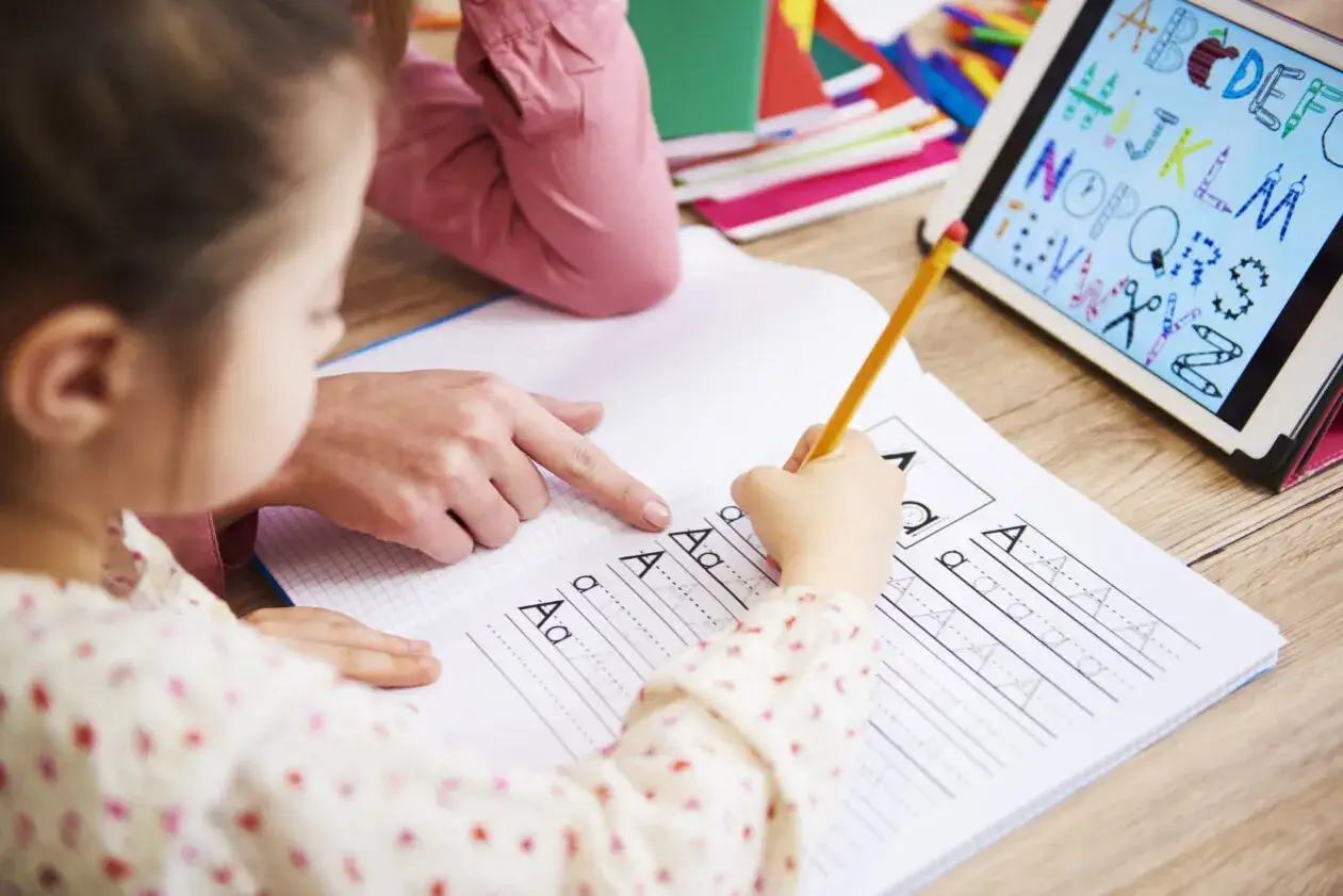 Dziecko ćwiczy pisanie liter, a dorosły pomaga. W tle tablet z alfabetem. To może być pierwszy krok do uzyskania zaświadczenia o dysleksji jak załatwić.